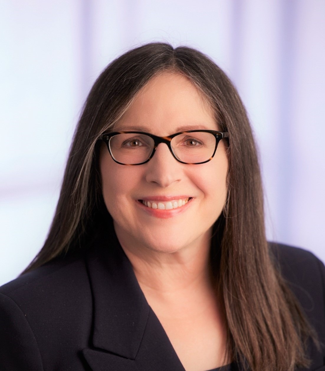 A smiling woman with long, straight hair and glasses wears a dark blazer. The soft background hints at a professional setting, suggesting expertise.